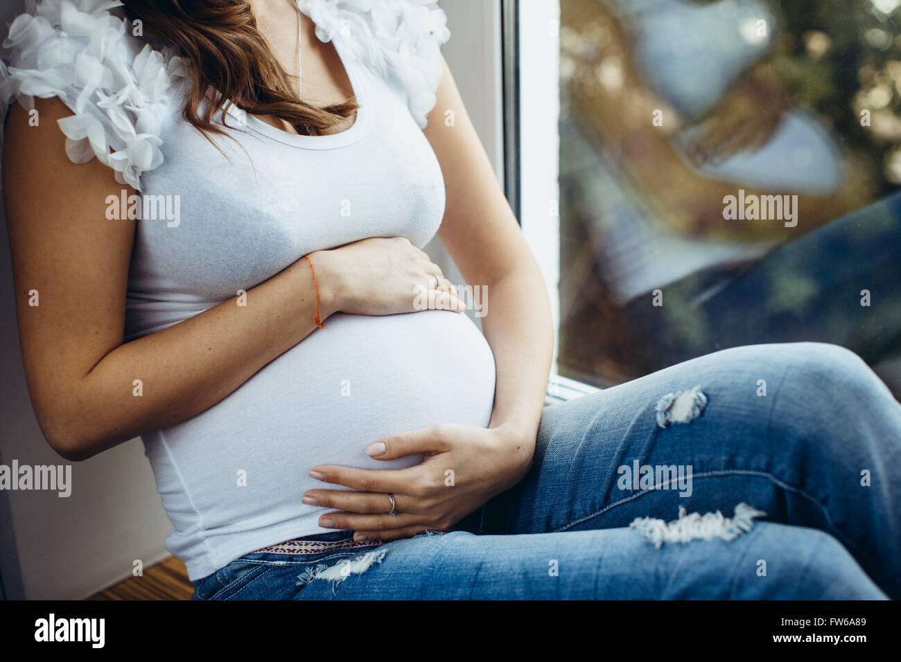 Belly closeup of a future mother. Pregnant brunette girl sitting on the windowsill, holding his stomach outside, the greens, the Stock Photo