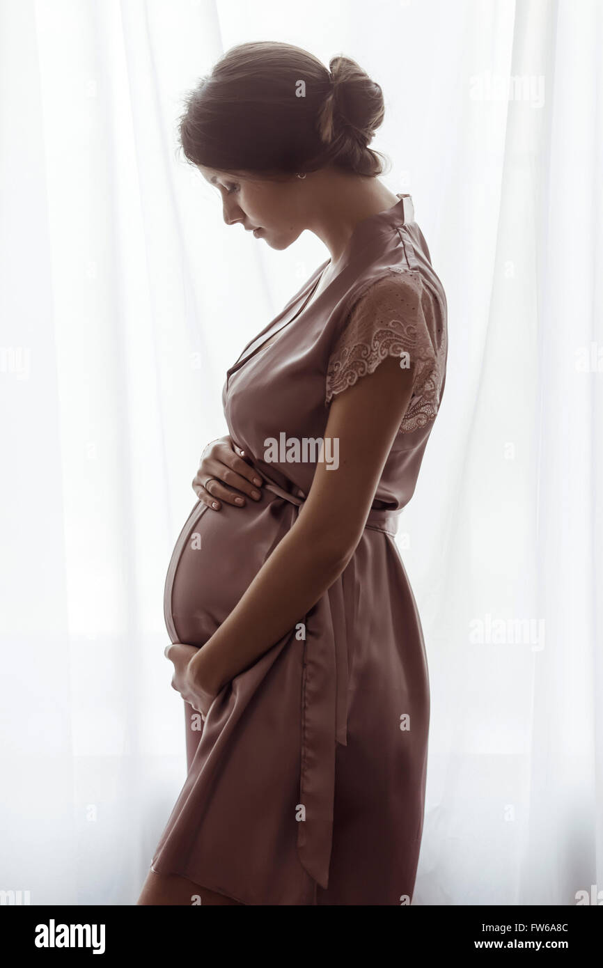 Silhouette of pregnant woman in a pink negligee standing against the window. Side view of happy pregnant woman looking at belly Stock Photo