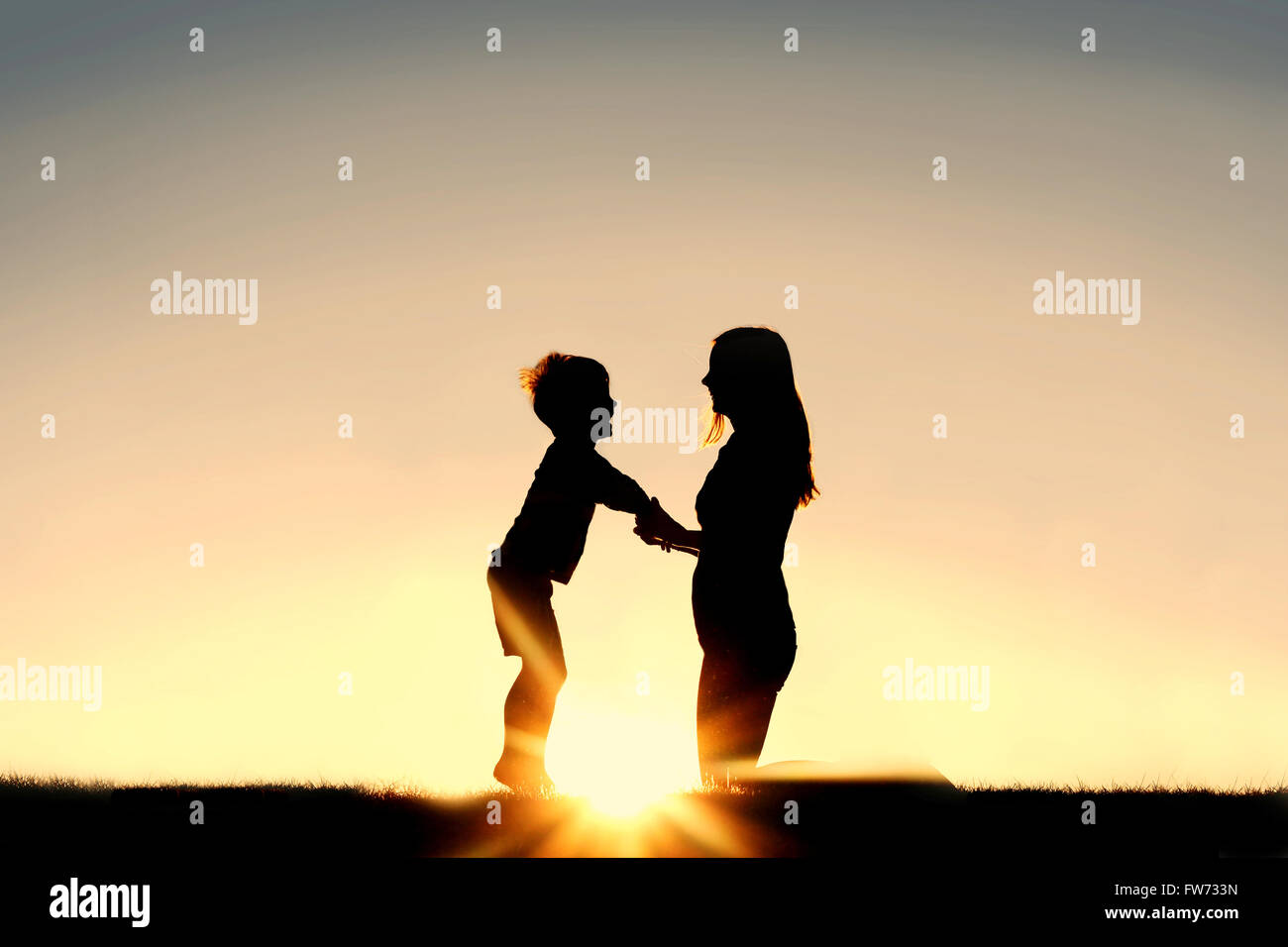 Silhouette of a young mother lovingly holding hands with her happy little child outside in front of a sunset in the sky. Stock Photo