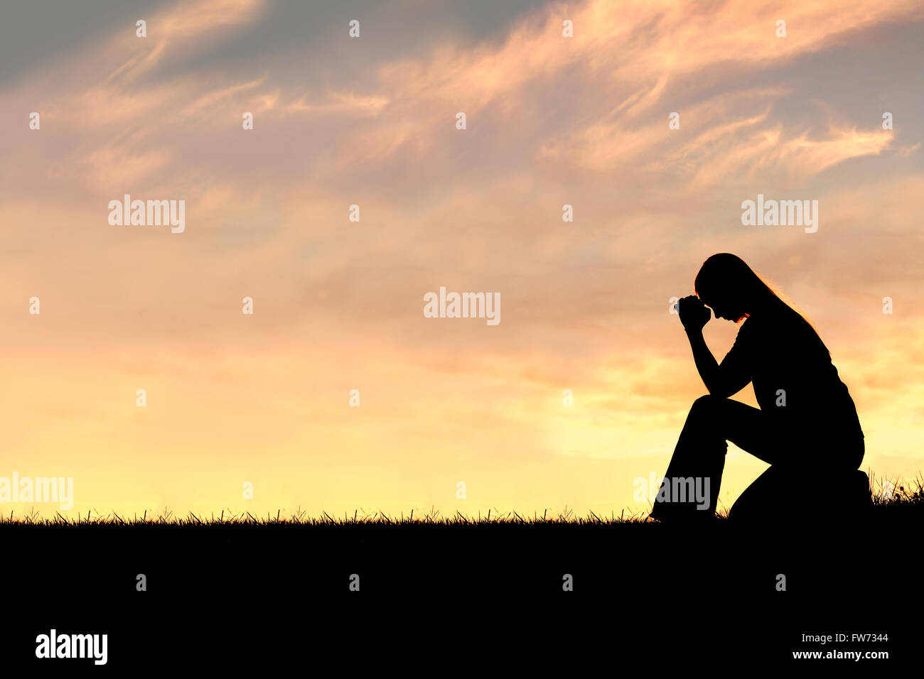 A silhouette of a young Christian woman is bowing her head in prayer, and desperation outside during sunset. Stock Photo