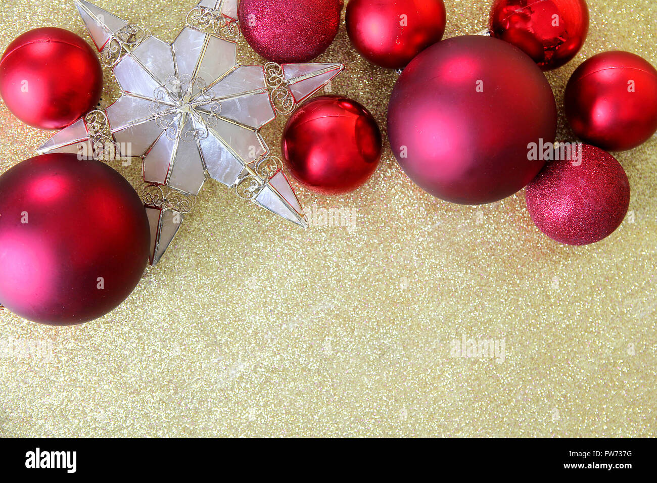 Red Christmas bulb decorations and a star shaped tree topper border the top of a background gold glitter fabric with copy-space. Stock Photo