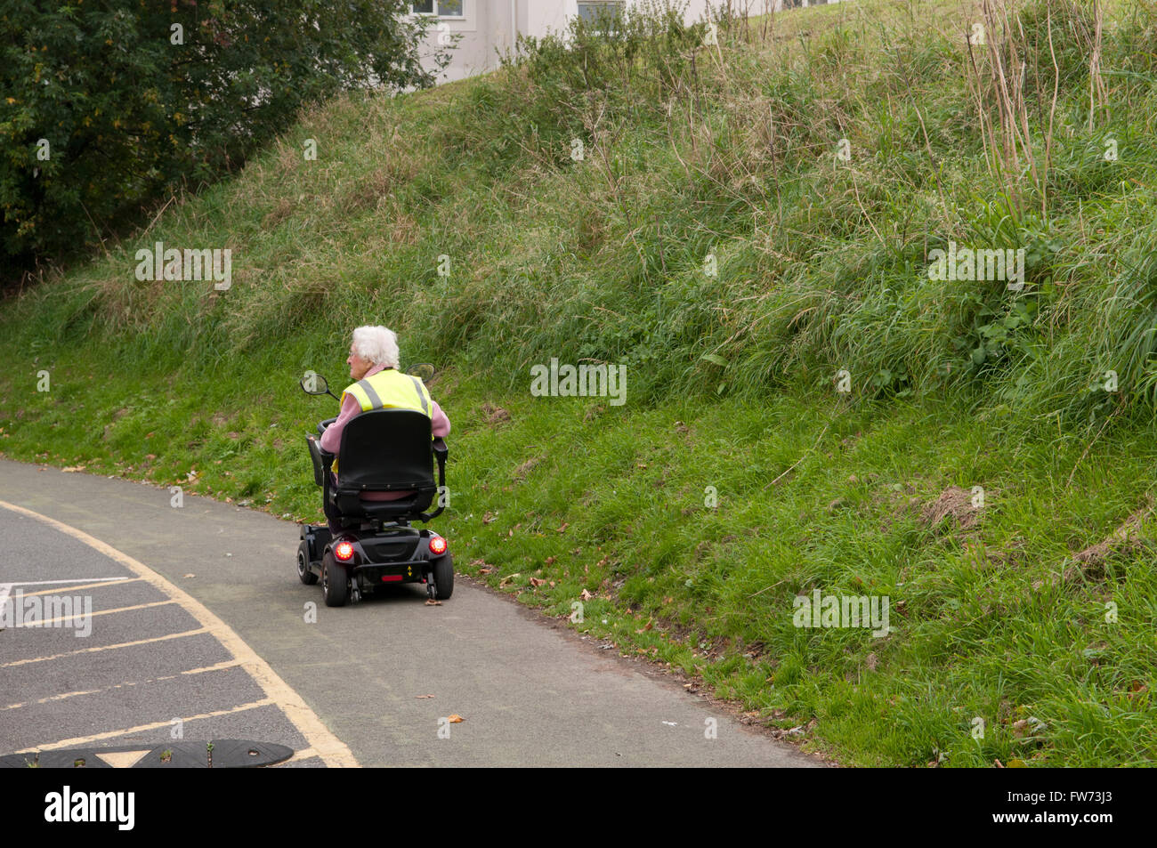 Rear view of an elderly woman using a mobility scooter wearing a yellow hi-vis safety vest Stock Photo