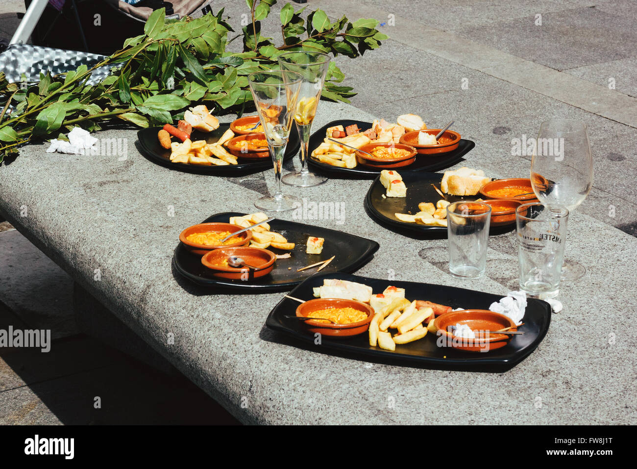 Several trays with food left on a stone bench. Tapas served in clay plates and green leaves in the background. Empty glasses Stock Photo