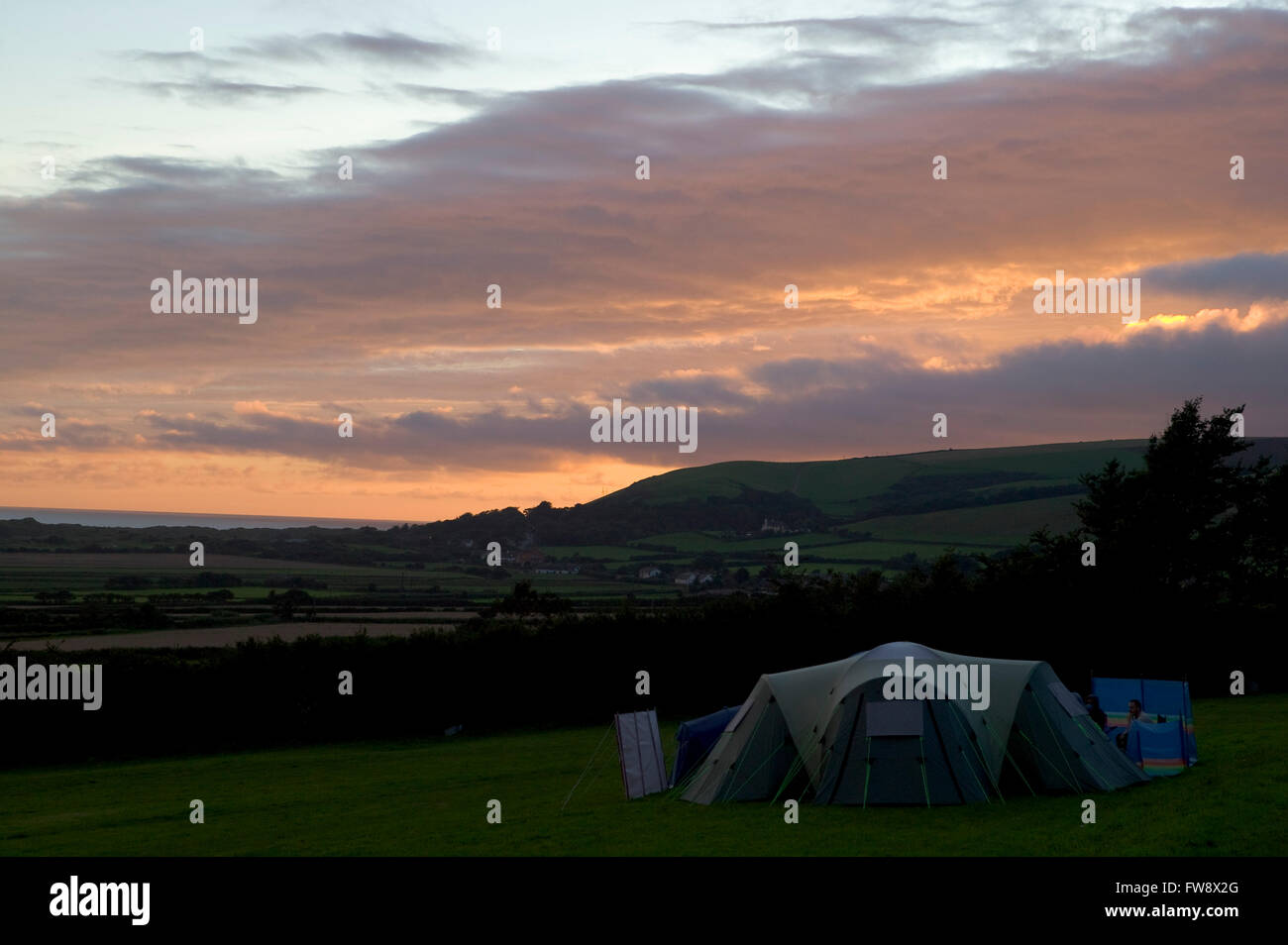The sunsets over the coast and in the foreground a tent is pitched in a field. The campers are gathered around the tent maybe eating an evening meal in this idylic summer holiday scene. Stock Photo