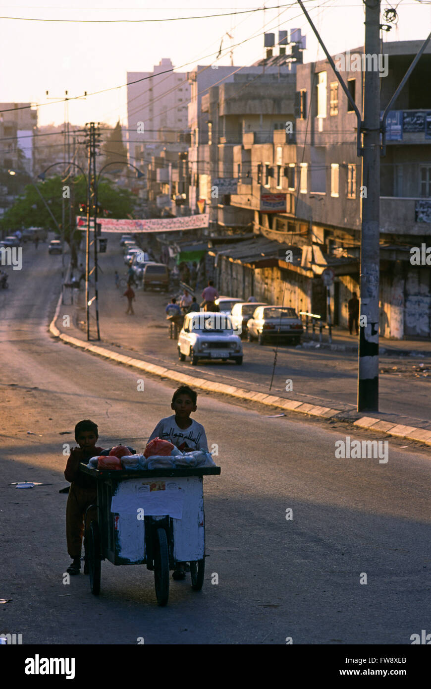 A street scene in Gaza city, Gaza strip contained on the borders of Israel in teh middle east. The scene shopws a typical sunset over a main street in the town with the road strewn with rubbish, old cars and people line the sidewlaks and pavements as the market begins to close down. Stock Photo