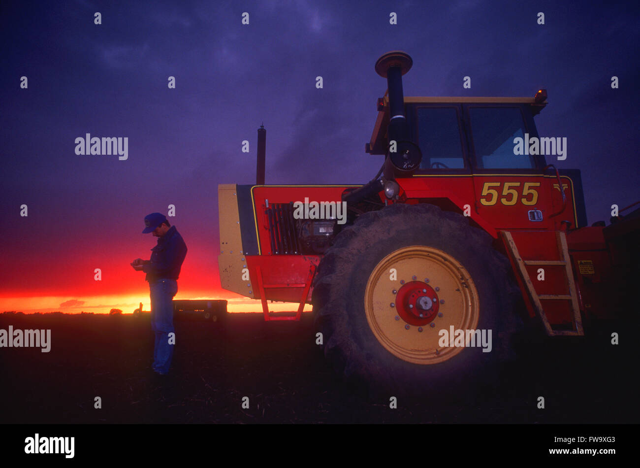 Farmer Standing In Front Of A Tractor In A Field At Sunset Stock Photo