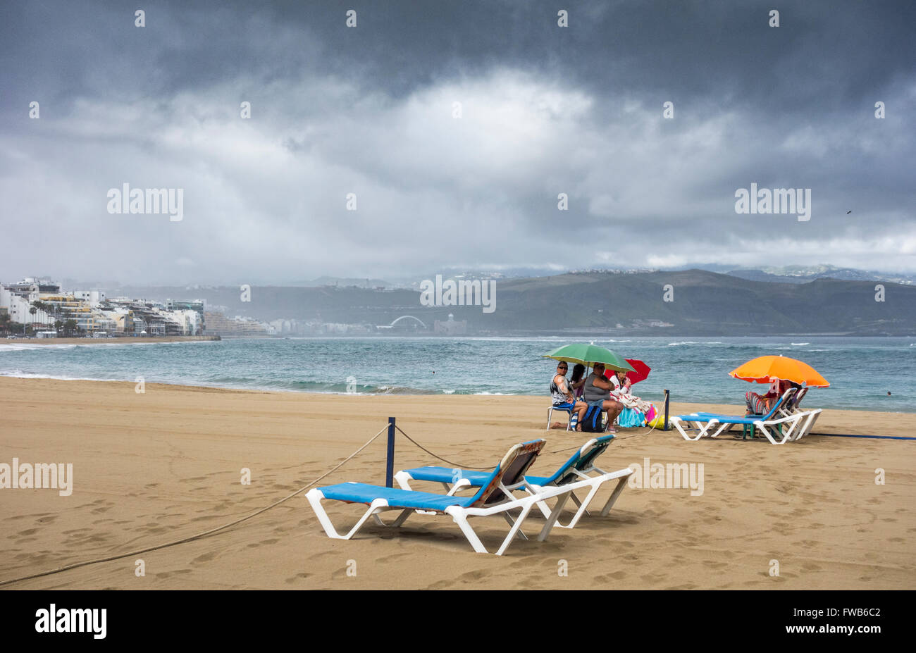 Las Palmas, Gran Canaria, Canary Islands, Spain, 3rd April 2016. Weather: With much of the UK forecast to have warm sunshine on Sunday and UK press saying 'hotter than Spain...', it`s a wet Sunday morning in Las Palmas on Gran Canaria as a band of rain sweeps over the city beach. PICTURED: A group of locals sit under parasols on Las Canteras beach waiting for Sunday morning rain to ease as black clouds pass overhead. Credit:  Alan Dawson News/Alamy Live News Stock Photo