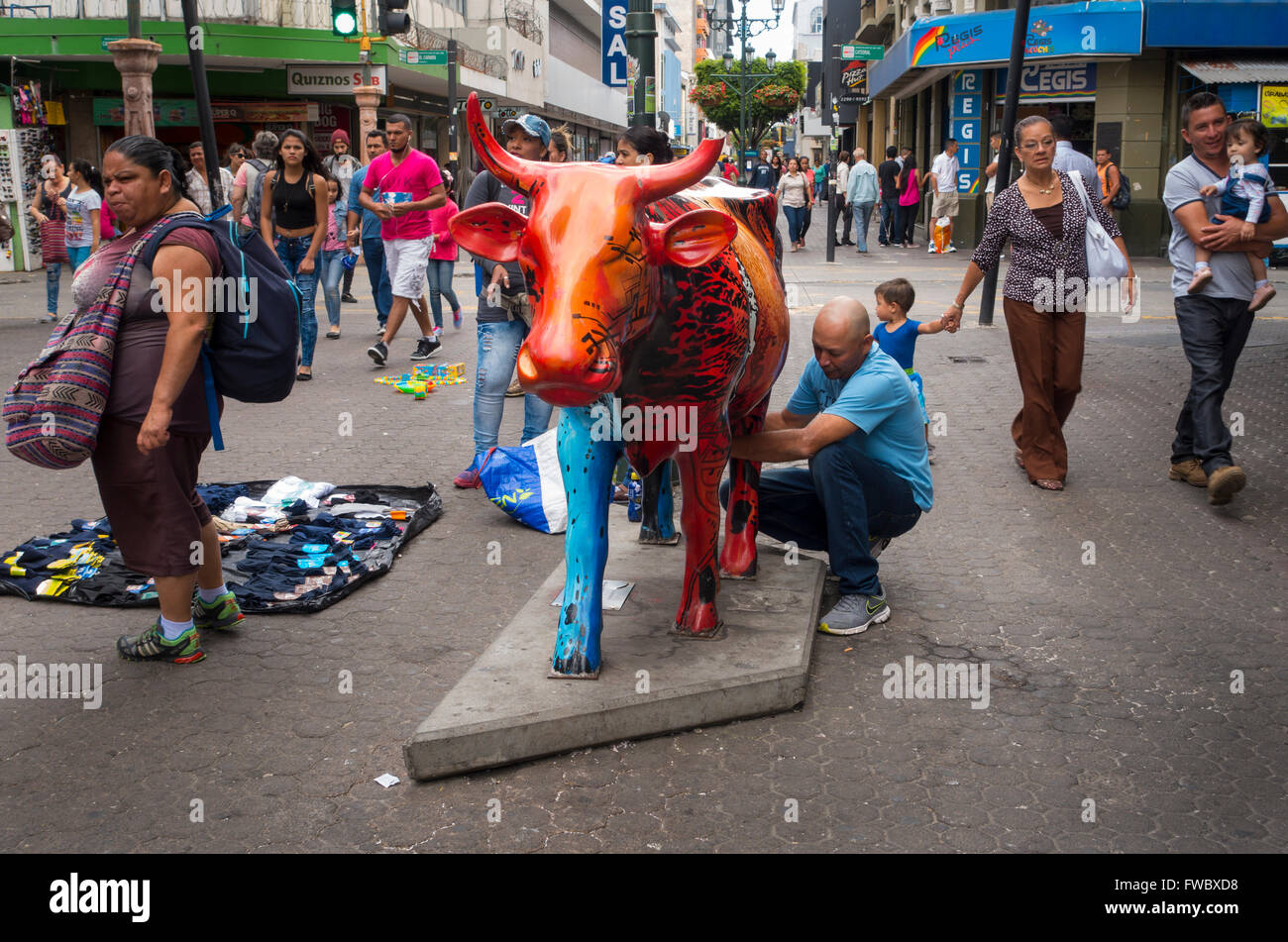 A tourist pretending to milk a cow statue. Avenida Central, San José, San José Province, Costa Rica. Stock Photo