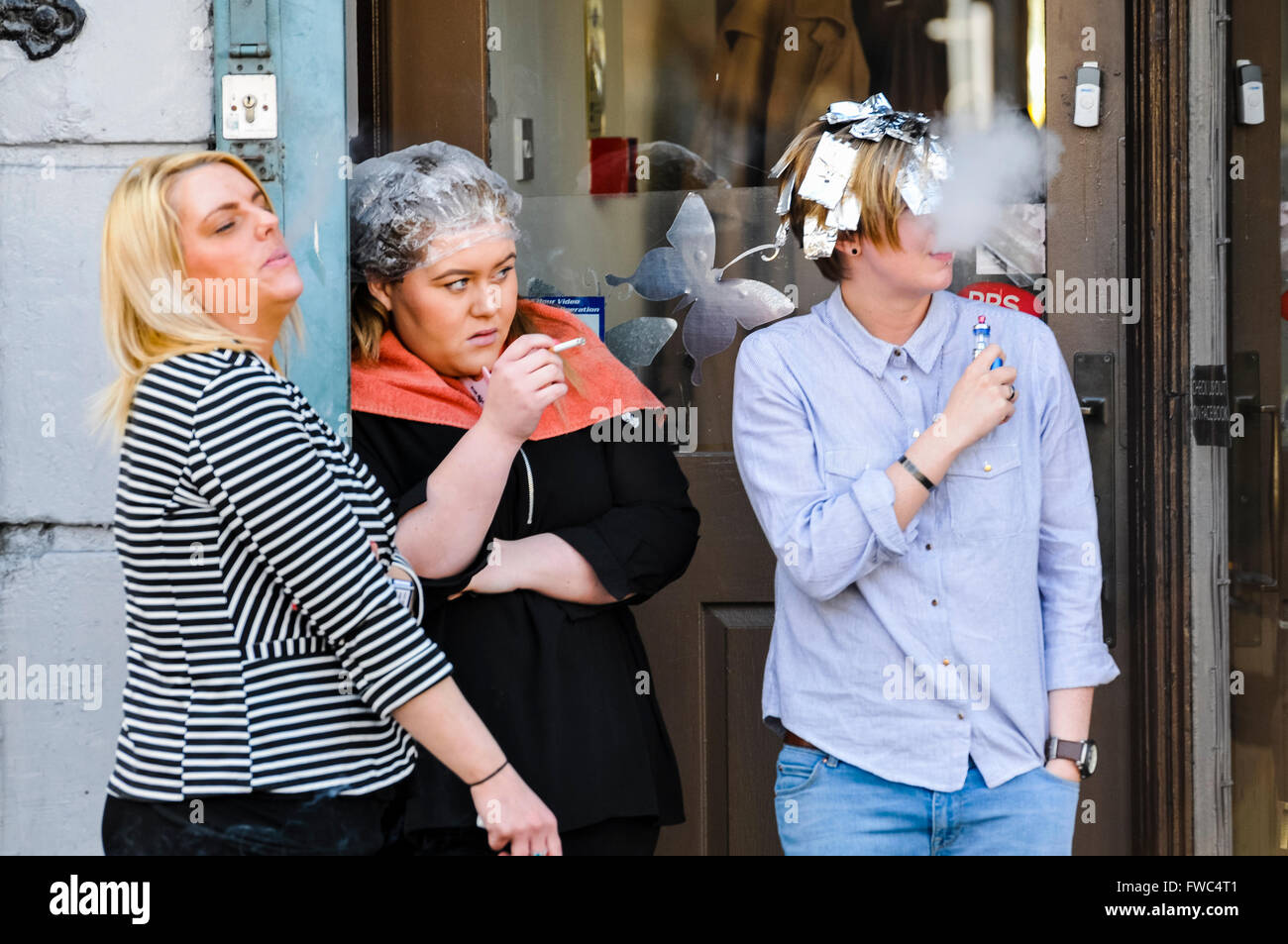 Three women stand at the doorway of a hairdressers shop, smoking and watching something happening. Stock Photo
