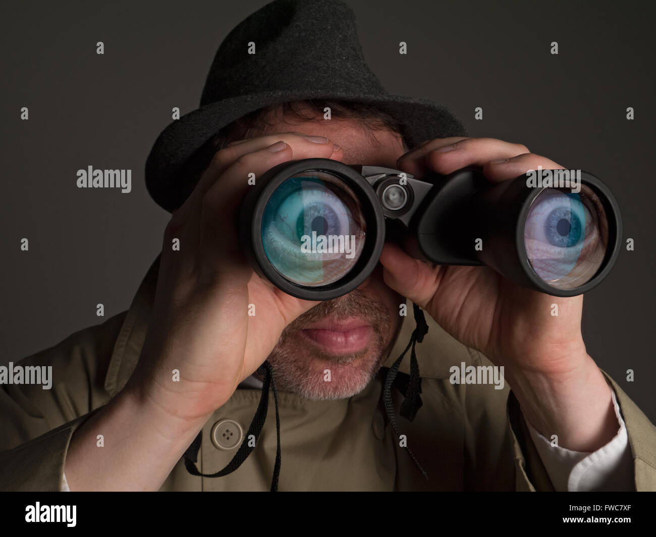 Photograph of a man in trench coat and hat looking through binoculars with huge, cartoonish eyes seen in the lenses. Stock Photo
