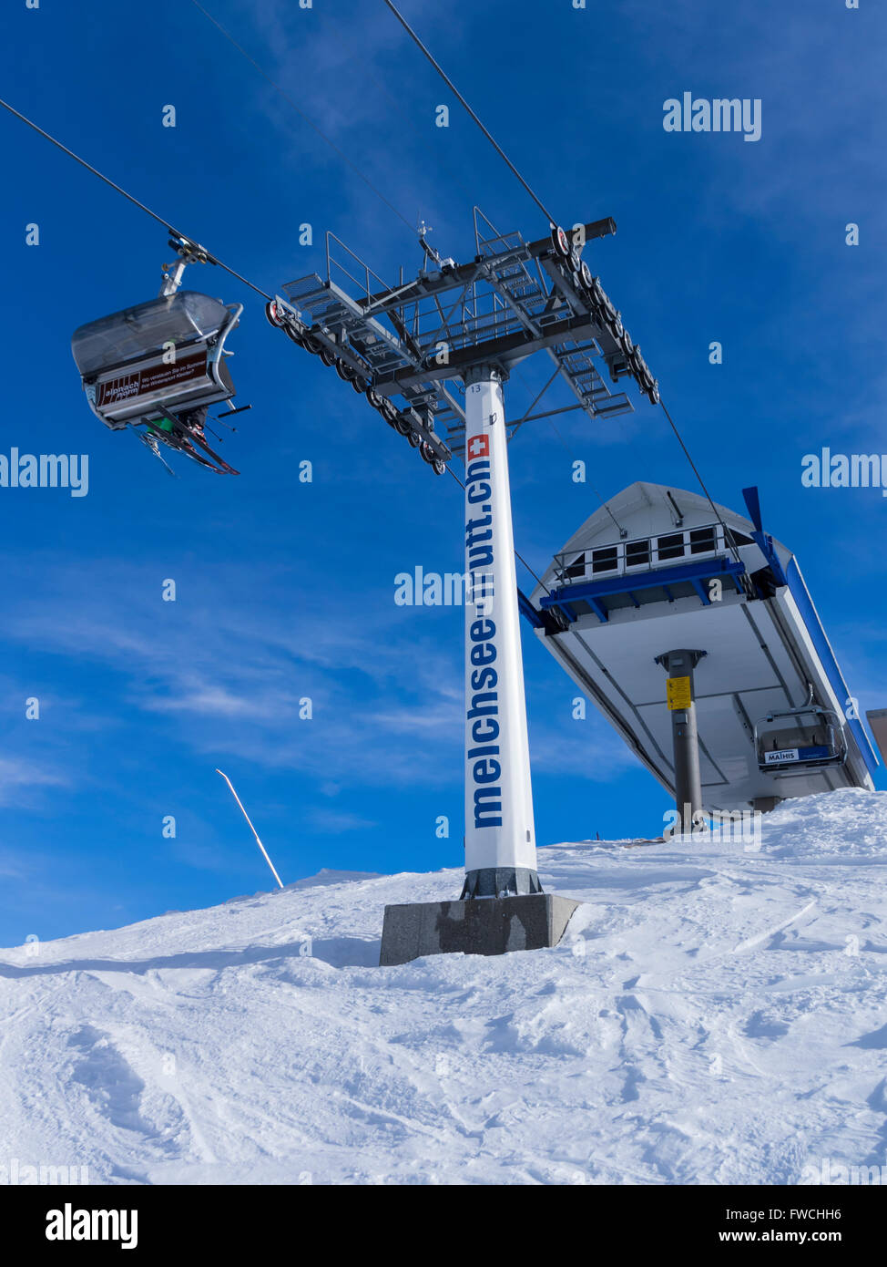 Chairlift in Melchsee-Frutt, Switzerland, with skiers on a chair, and a mast advertising the URL of the winter sports resort. Stock Photo