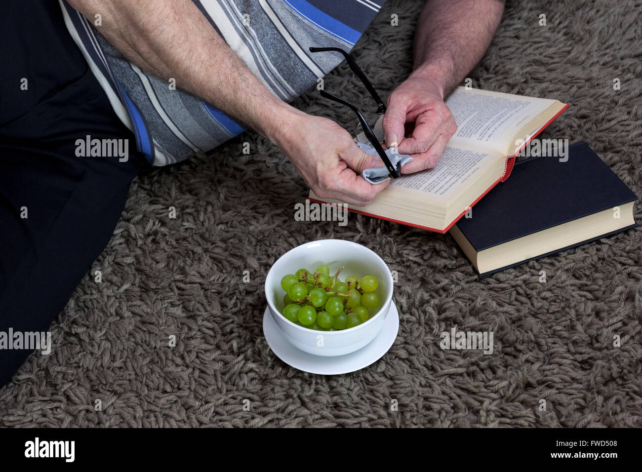 Man laying on a shaggy grey rug cleaning his glasses while reading a red book Stock Photo
