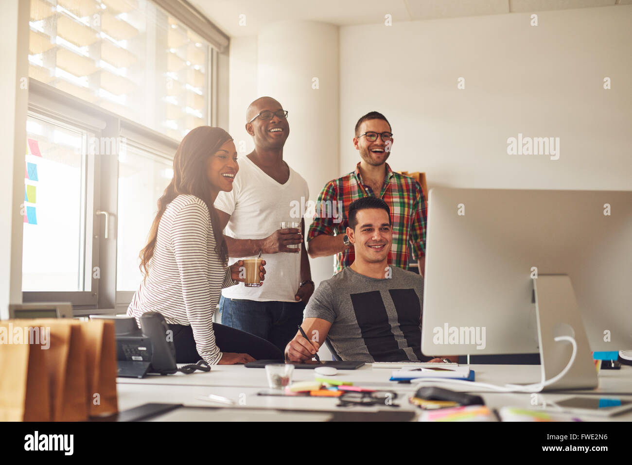 Casually dressed laughing woman and three men holding drinks around computer with stylus and tablet on desk in office Stock Photo