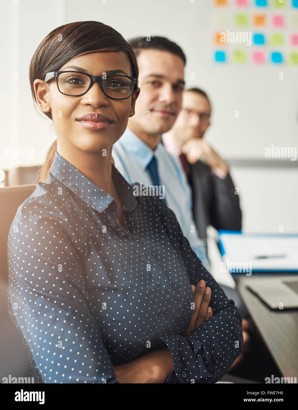 Confident smiling business woman wearing eyeglasses and white polka dotted shirt seated with folded arms beside male co-workers Stock Photo