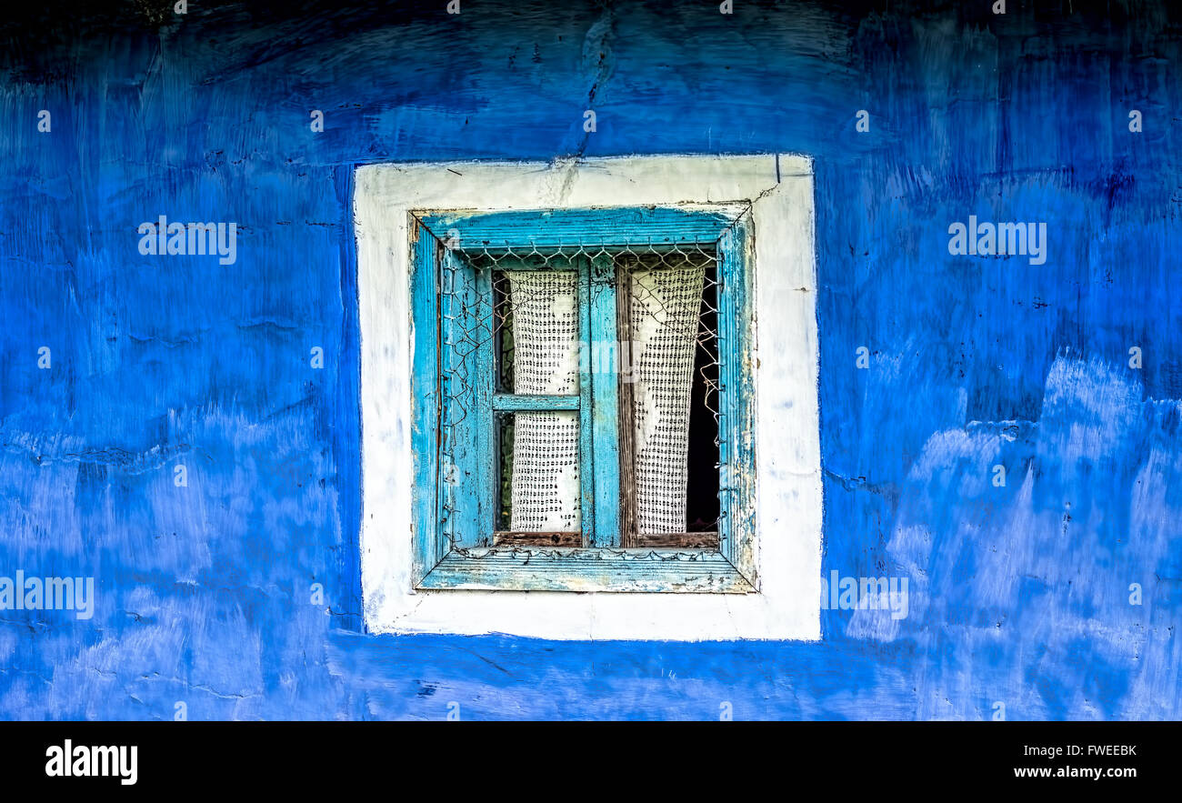 Old window with cyan wooden frame  and crocheted curtain on a blue plastered hand painted wall. Stock Photo