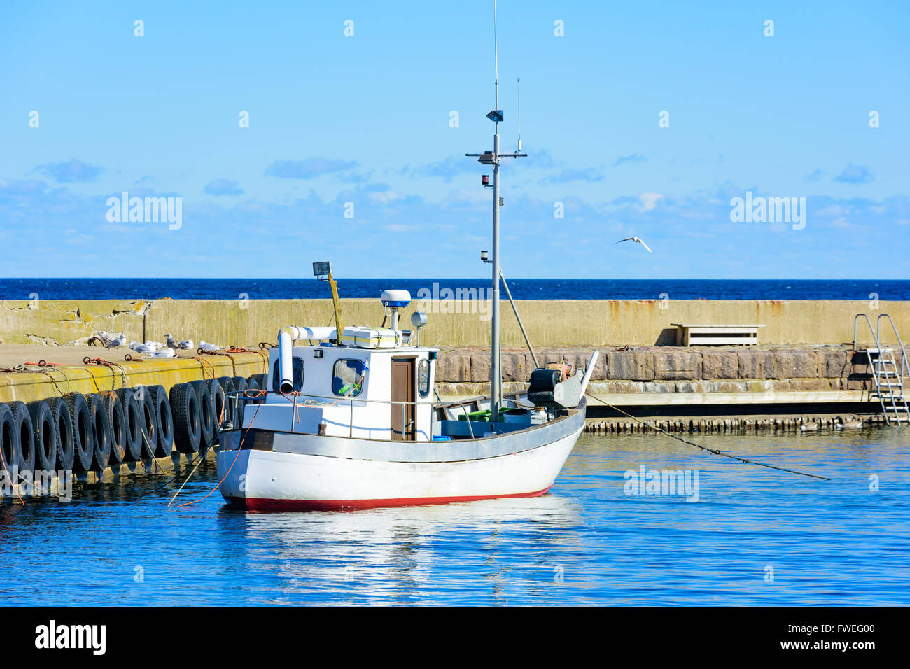 Small white fishing boat moored by the stone pier in the harbor. Gulls sit beside it on the pier. Fine, sunny weather and very l Stock Photo