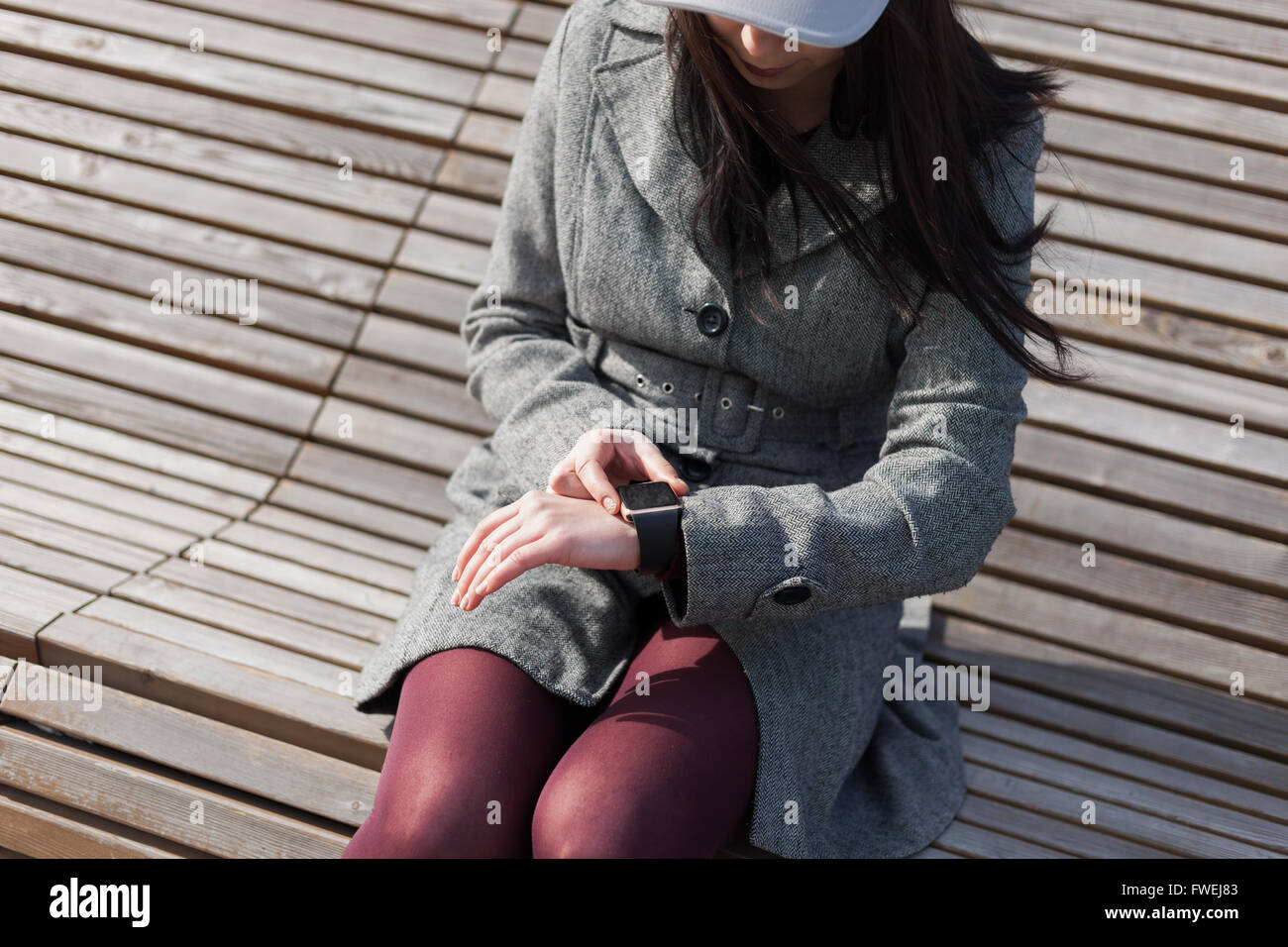 Young brunette girl in grey coat using her smart watch while sitting on the bench outdoors. This person is always connected to social media and internet everywhere. Stock Photo