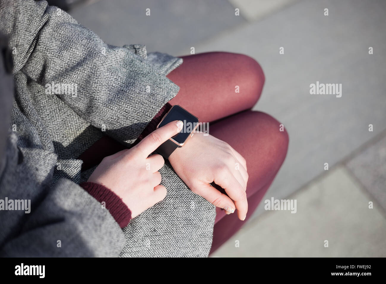 Woman in grey coat and marsala tights using her trendy smart wrist watch. This person is always connected to social media and internet. Stock Photo