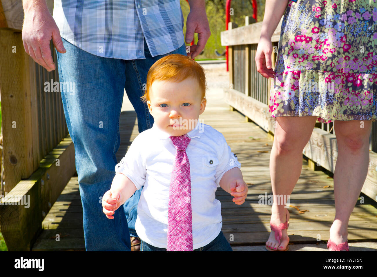 A baby wearing a red or pink necktie walks across a bridge with his parents behind him. Stock Photo