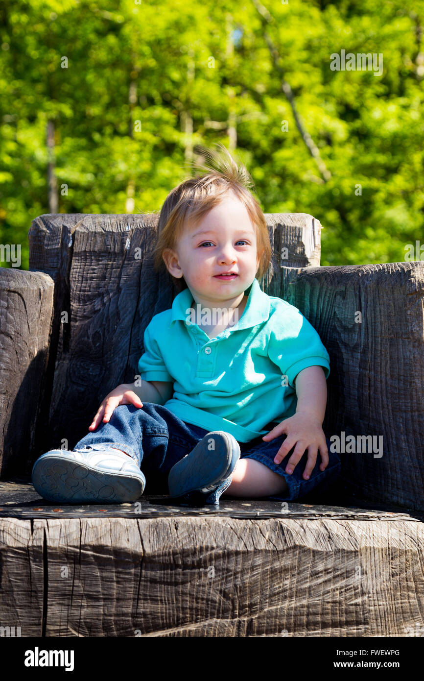 A cute young boy sits on a stump carved into a seat at a park outdoors for this cute and simple portrait. Stock Photo