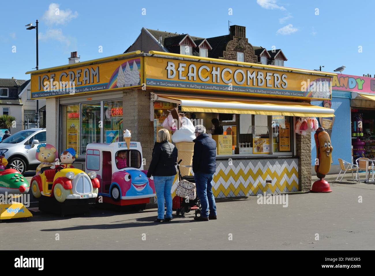 Parents watch their child on a child's amusement ride, on the Largs seaside next to a promenade shop, in Scotland, UK. Stock Photo