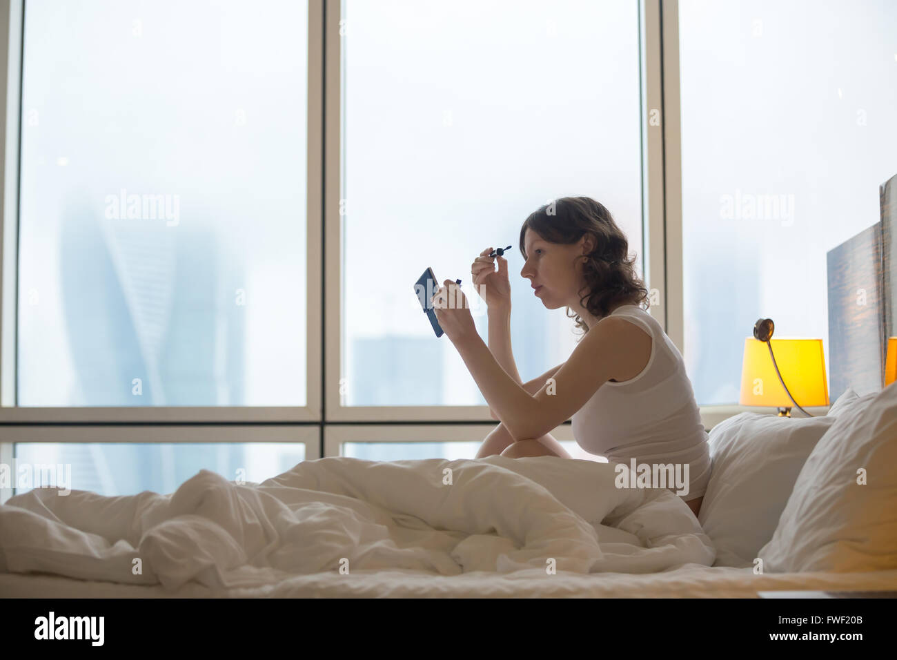 Portrait of young woman sitting on unmade bed after waking up and putting on mascara. Caucasian female model Stock Photo