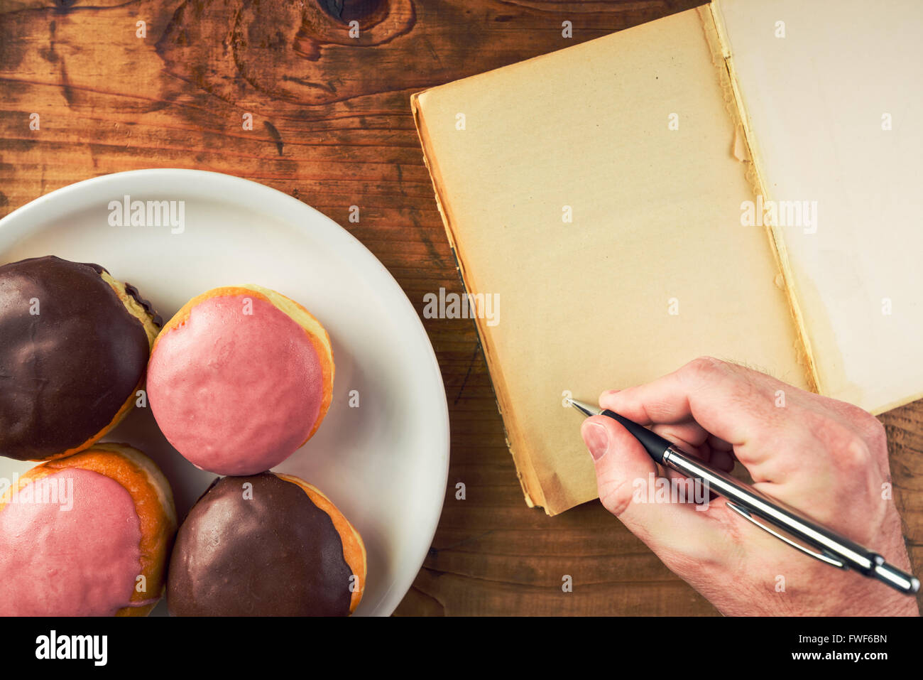 Writing recipe for homemade delicious doughnuts with sweet topping, top view of male hand writing in notebook and tasty donuts Stock Photo