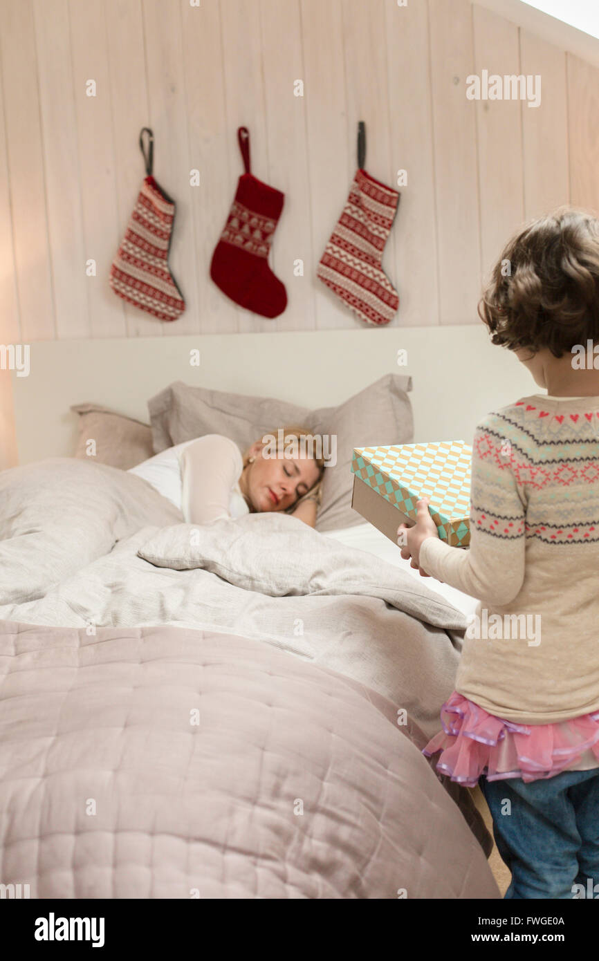 A woman in bed on Christmas morning, and a young girl standing beside her carrying presents. Stock Photo