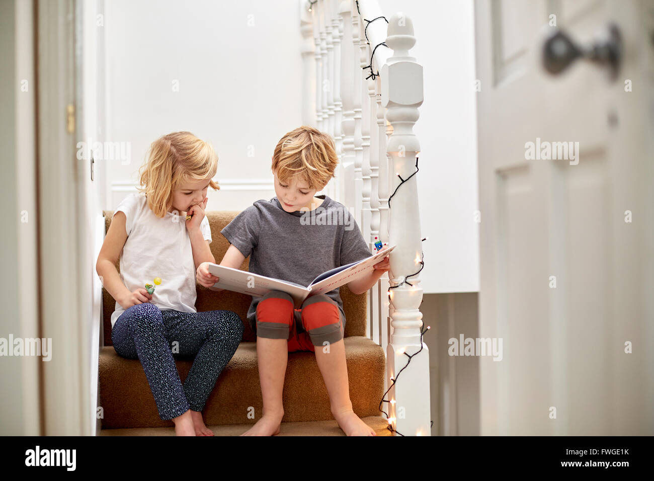 Two children seated side by side on the stairs at home, reading a book together. Stock Photo