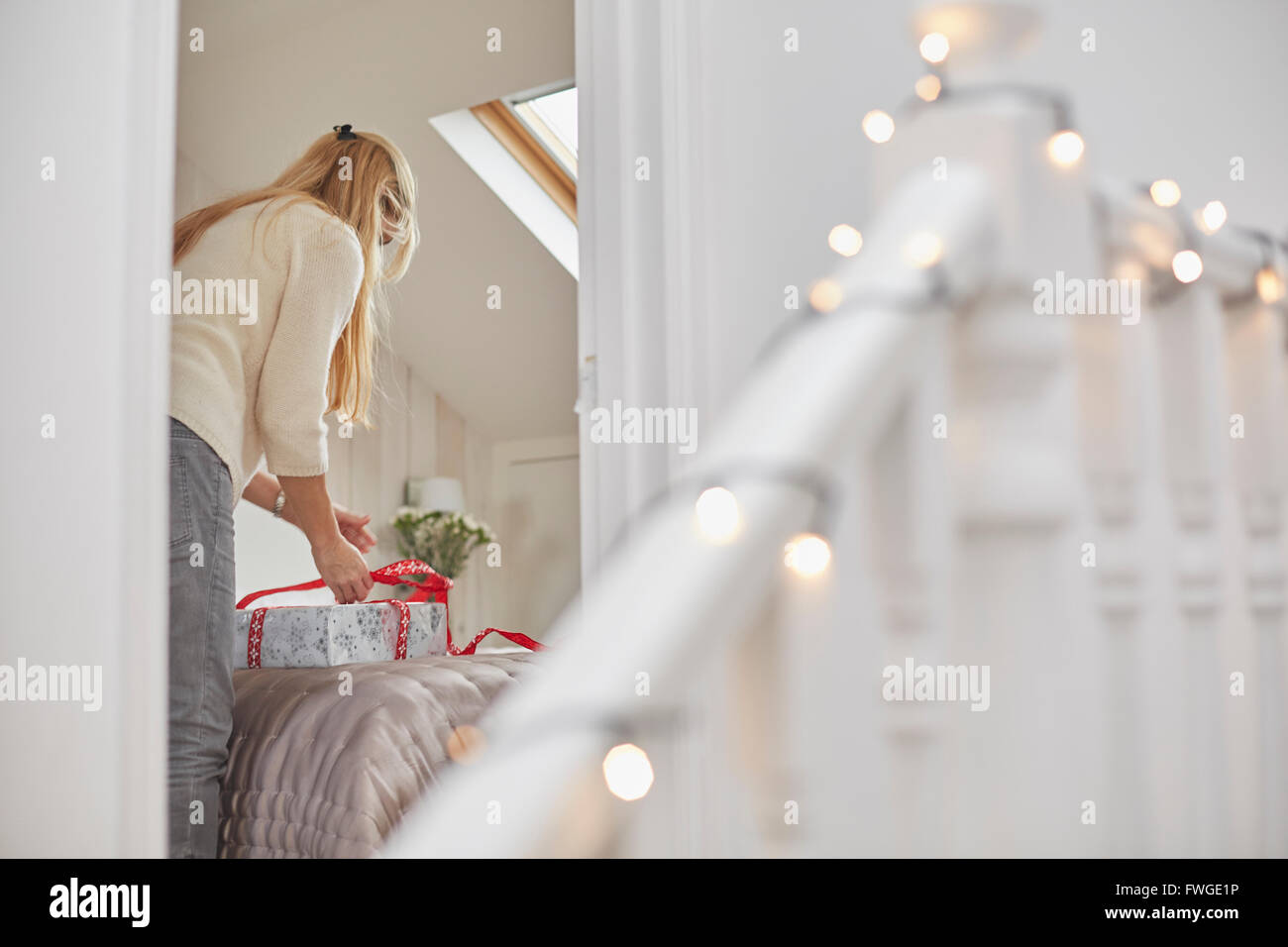 A woman wrapping red ribbons around Christmas presents on a bed, view through the bedroom door. Stock Photo