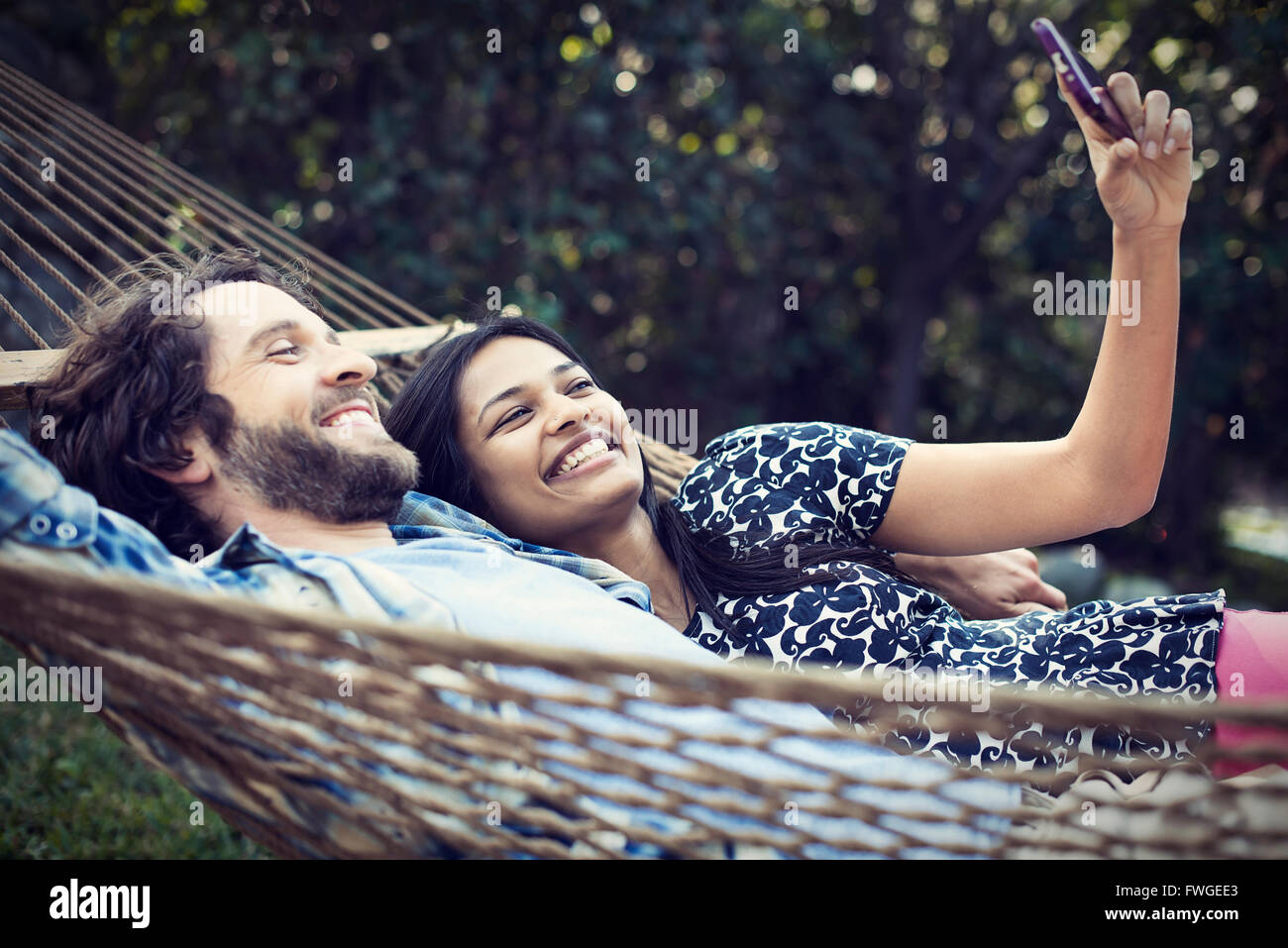 A couple, a young man and woman lying in a large hammock in the garden, taking a selfy of themselves. Stock Photo