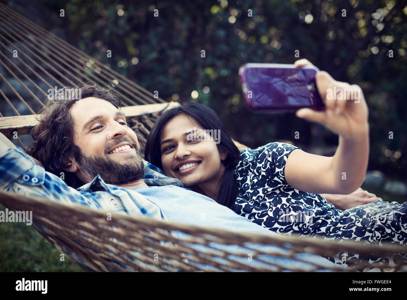 A couple, a young man and woman lying in a large hammock in the garden, taking a selfy of themselves. Stock Photo
