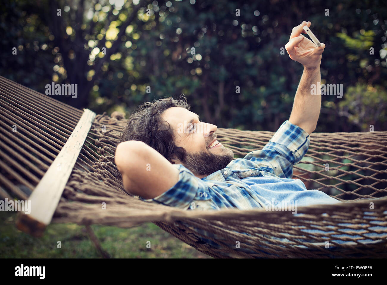 A man lying in a garden hammock taking selfies with his phone. Stock Photo