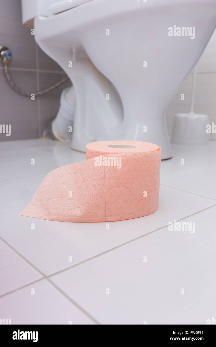 Roll of soft absorbent pink toilet paper on the clean white tiled floor of a bathroom at the foot of the toilet bowl, low angle Stock Photo