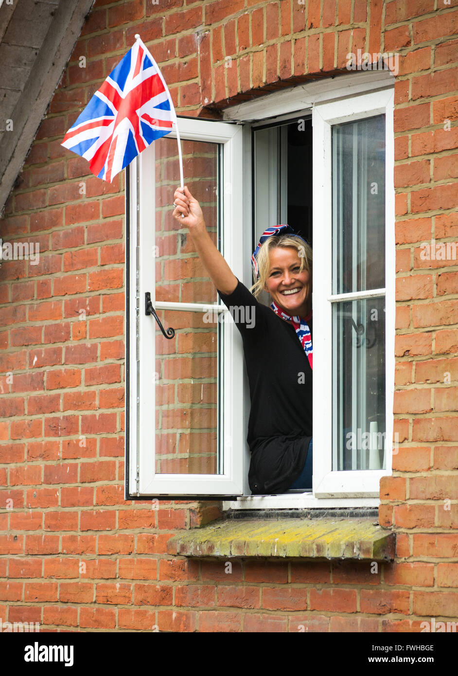 Happy, smiling woman waving a Union Jack flag from a window. Stock Photo
