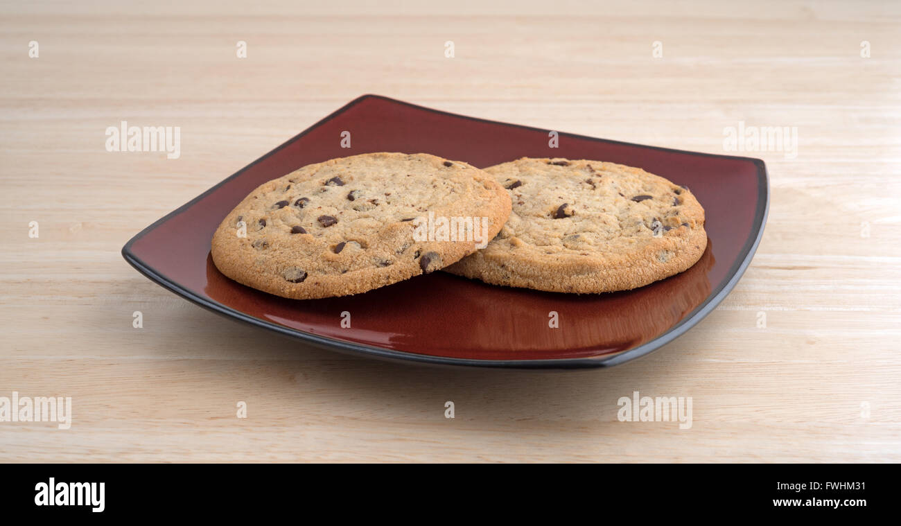 Two chocolate chip gluten-free cookies on a deep red plate atop a wood table top. Stock Photo