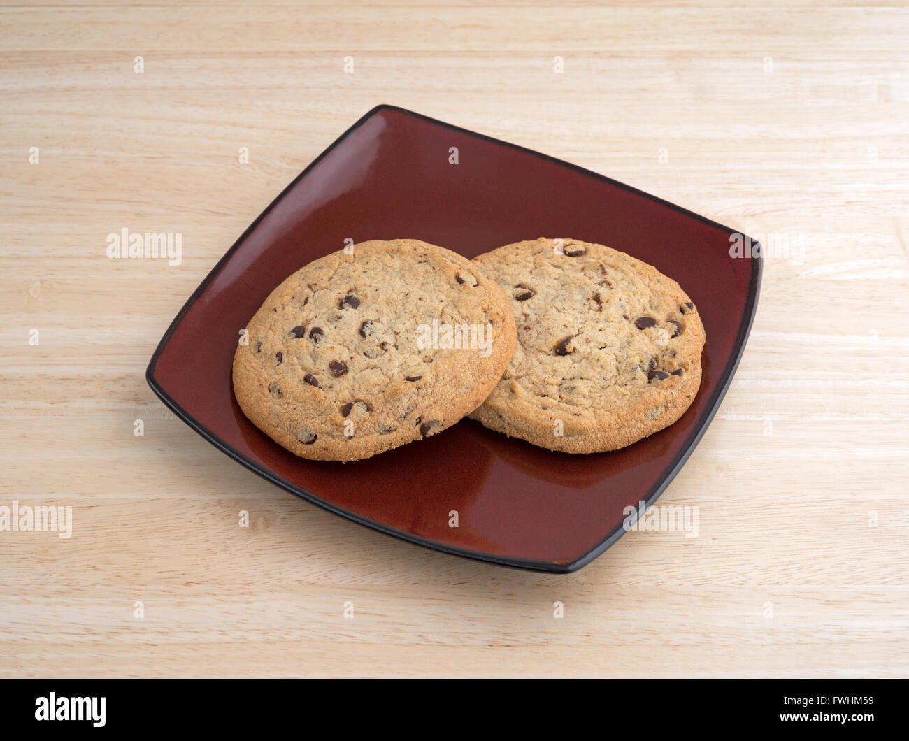 Two chocolate chip gluten-free cookies on a deep red plate atop a wood table top. Stock Photo