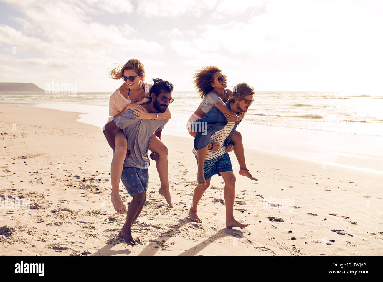 Two beautiful young couples walking along the beach, with men giving piggyback ride to women. Piggyback games on beach vacation. Stock Photo
