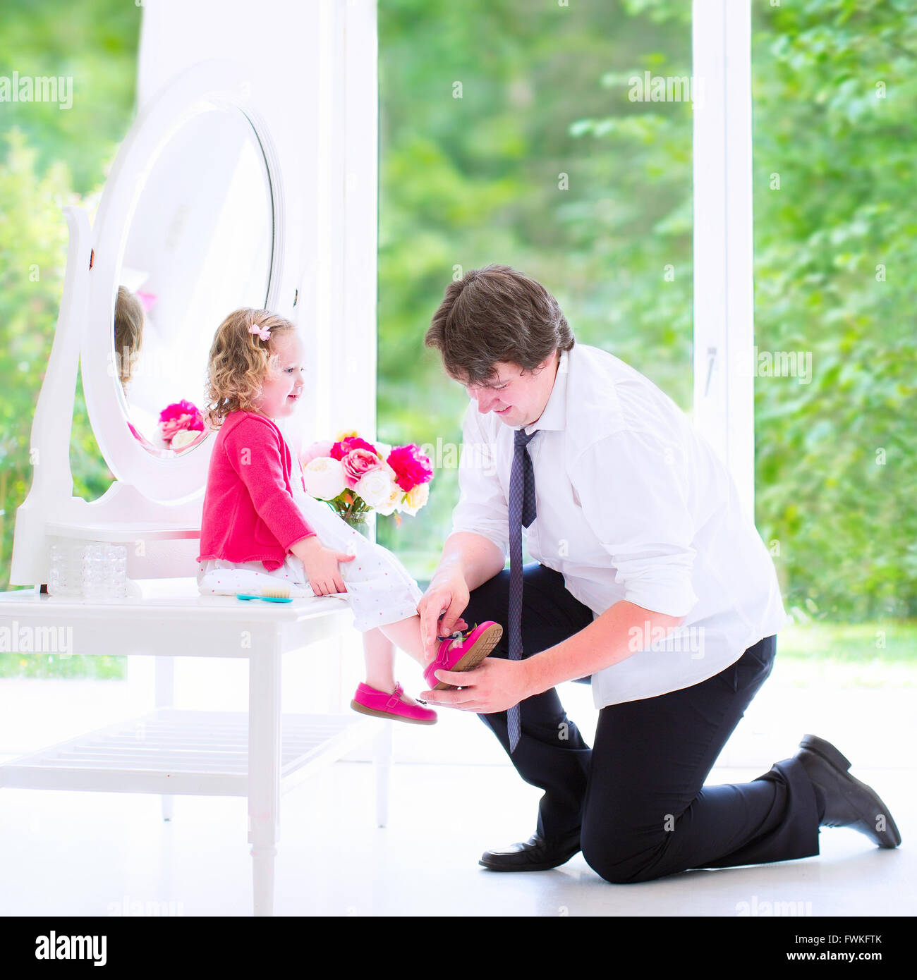 Young happy father in a business suit and tie helping his little daughter, cute curly toddler girl to put on a shoe Stock Photo