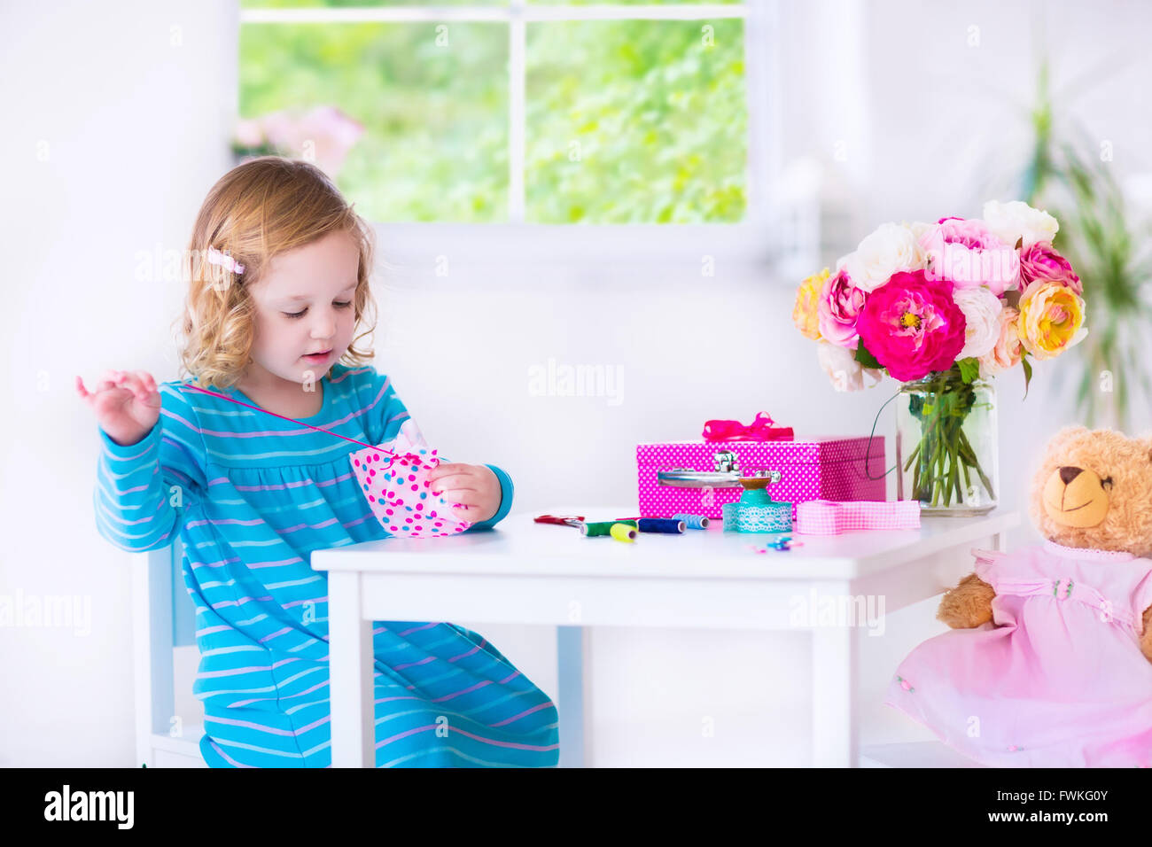 Cute creative little girl sewing a dress for her teddy bear doll, playing with needles and ribbons in white sunny room at home Stock Photo