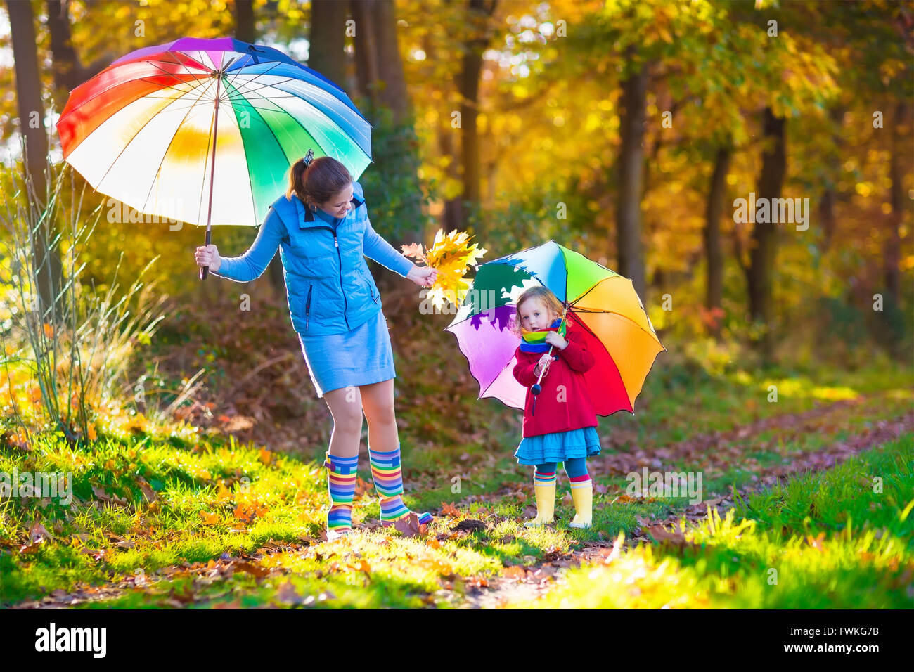 Mother and daughter play in autumn park with golden leaves. Child holding umbrella in the rain. Parent and kid walk in forest Stock Photo