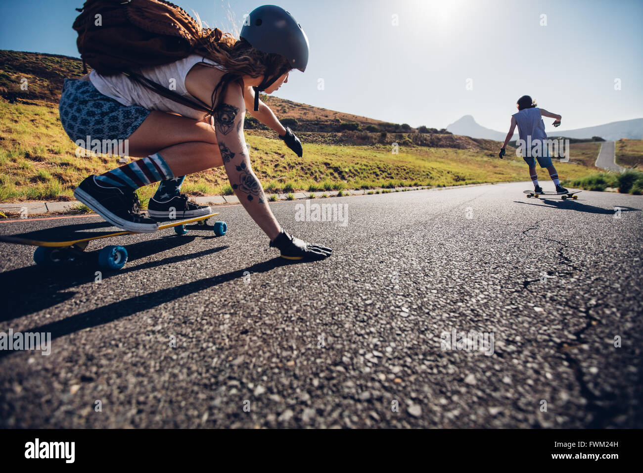 Young woman skating with her friend in background. Young people longboarding on rural road on summer day. Stock Photo