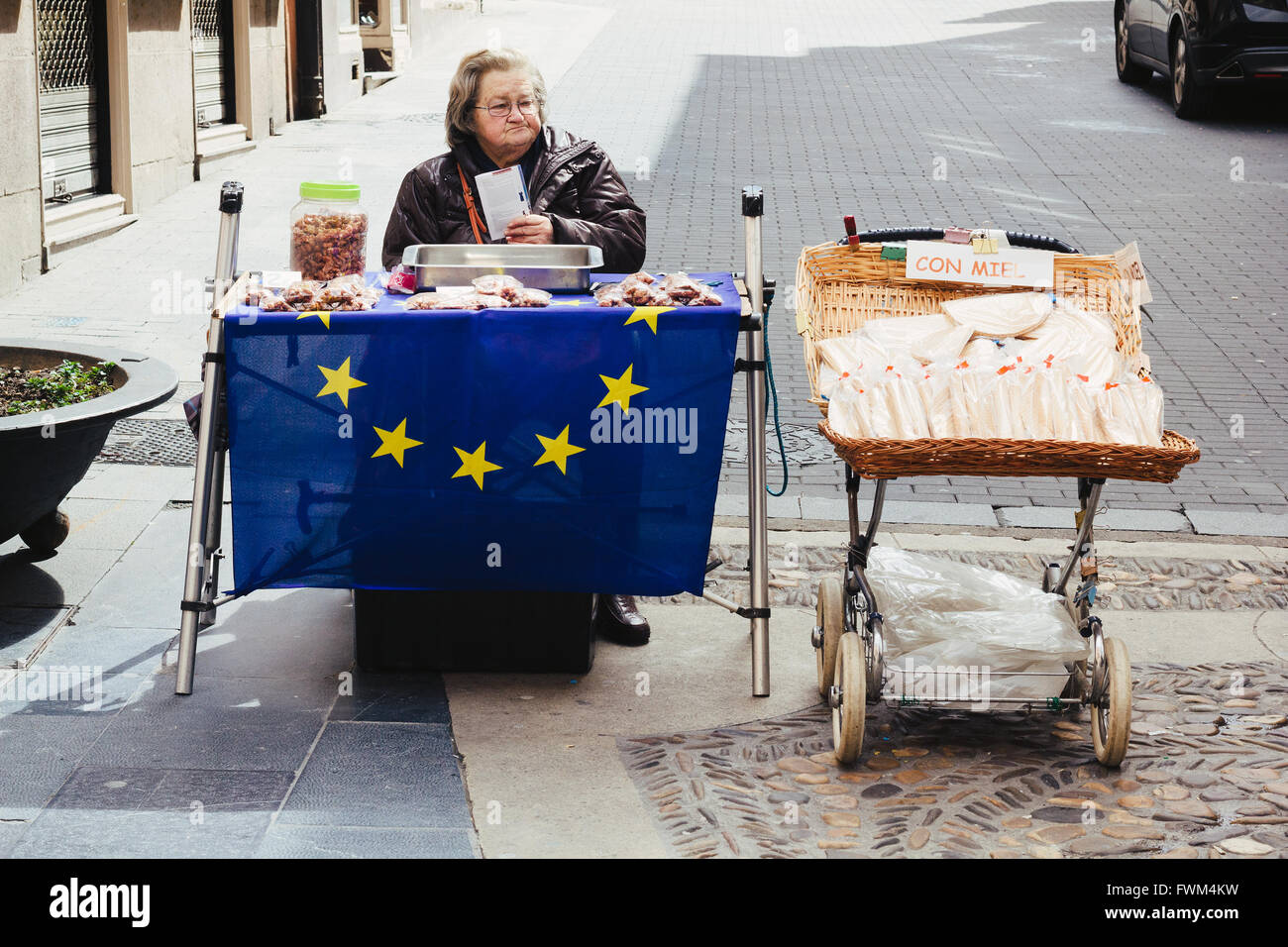 Old woman outdoors, sitting down, looking sideways, selling traditional sweets on a stall covered by the European flag. Spain Stock Photo