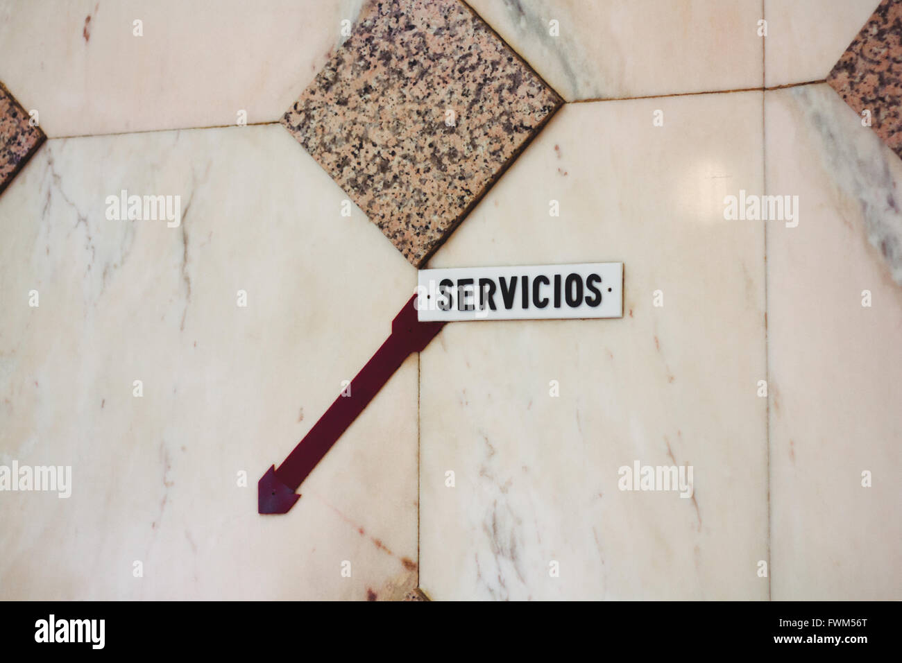 Toilet sign written in Spanish with a dark red arrow pointing downstairs on a marble wall. Stock Photo