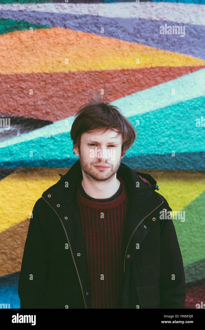 Young caucasian man looking at the camera, wearing warm/casual clothes.  Colour urban wall in the background. Stock Photo