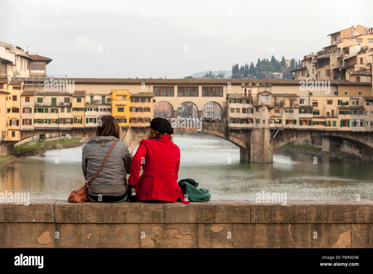 A couple sit on a bridge looking at the Ponte Vecchio, in Florence, Italy. Stock Photo
