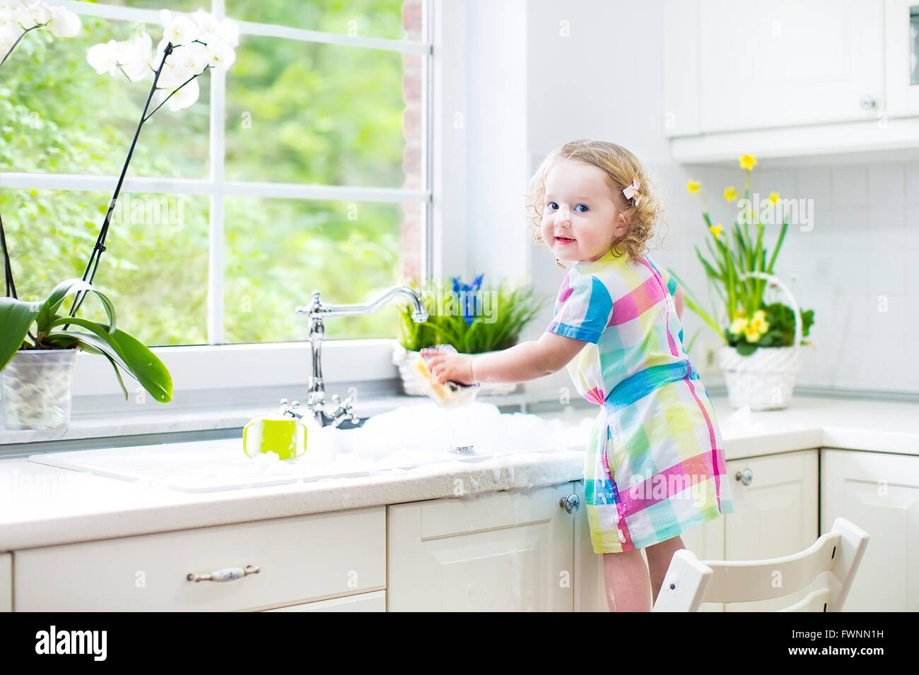 Cute curly toddler girl in a colorful dress washing dishes, cleaning with a sponge and playing with foam in the sink Stock Photo