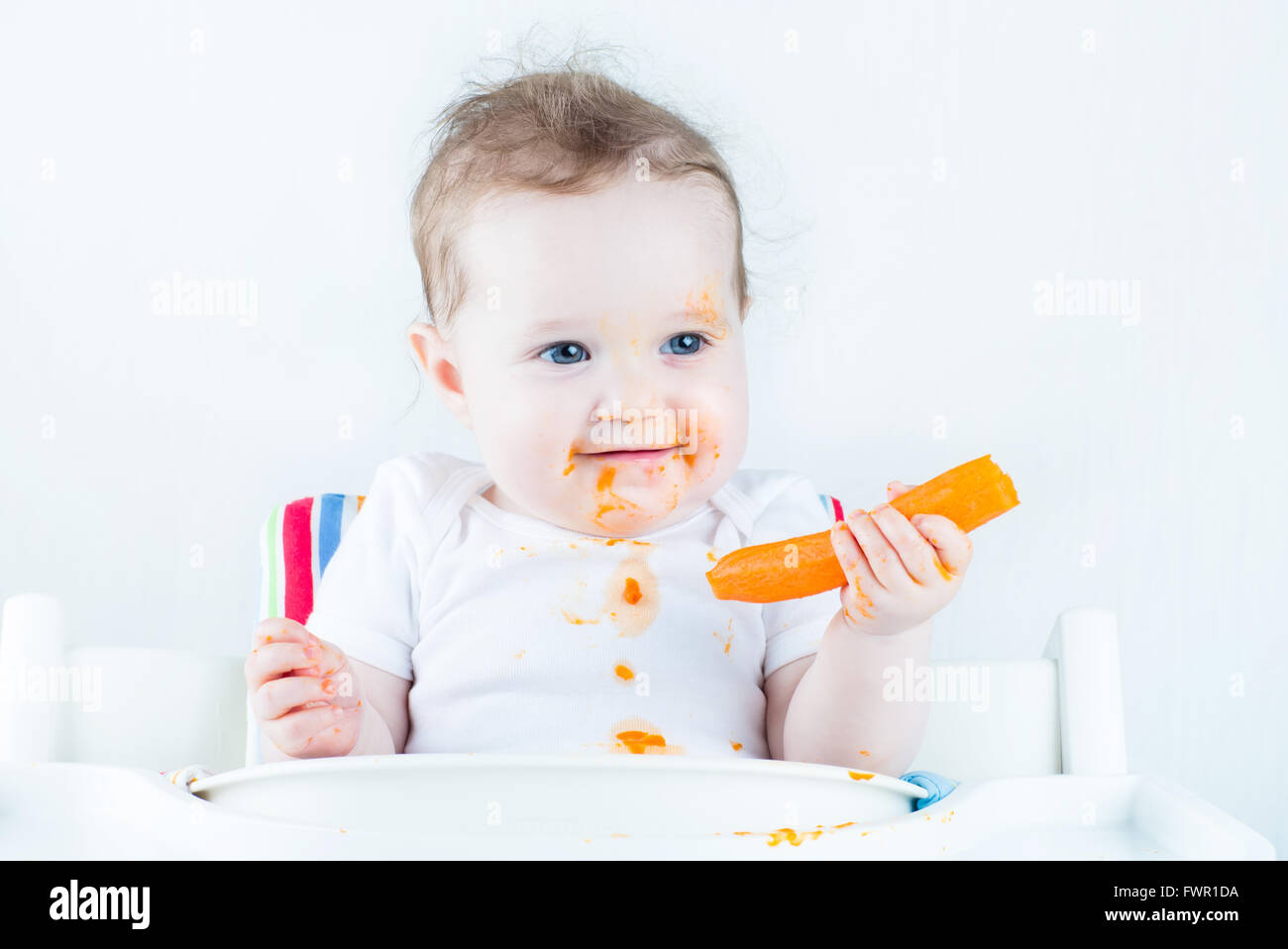 Funny laughing baby girl eating a carrot trying her first solid vegetable food sitting in a white high chair, isolated on white Stock Photo