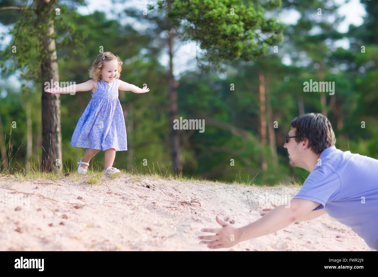 Little curly happy girl playing with her father,  running from a sand dune in a pine wood forest on a sunny warm summer day Stock Photo