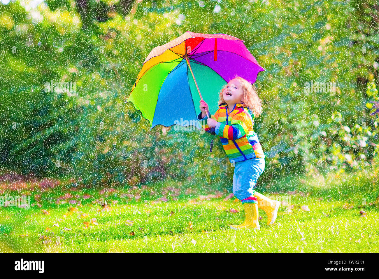 Funny cute curly toddler girl wearing yellow waterproof coat and boots holding colorful umbrella playing in the garden Stock Photo
